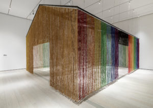 Long strands of various colors form a structure that hangs in an all-white room.