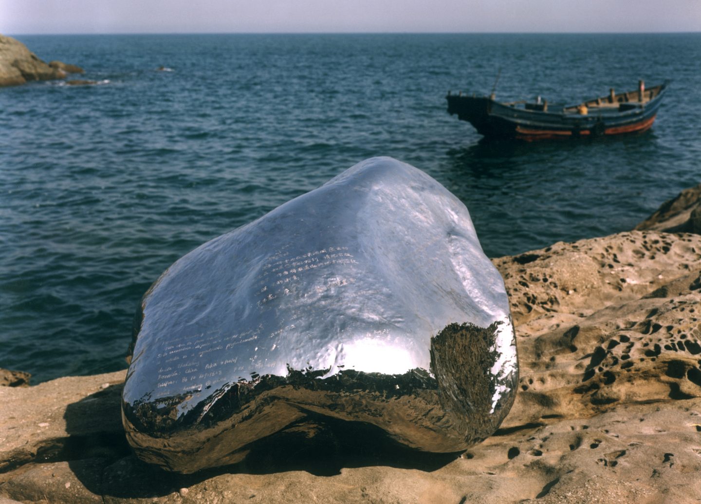 A large, shiny silver rock-like object rests on a rocky shoreline. A small boat floats in the water nearby.